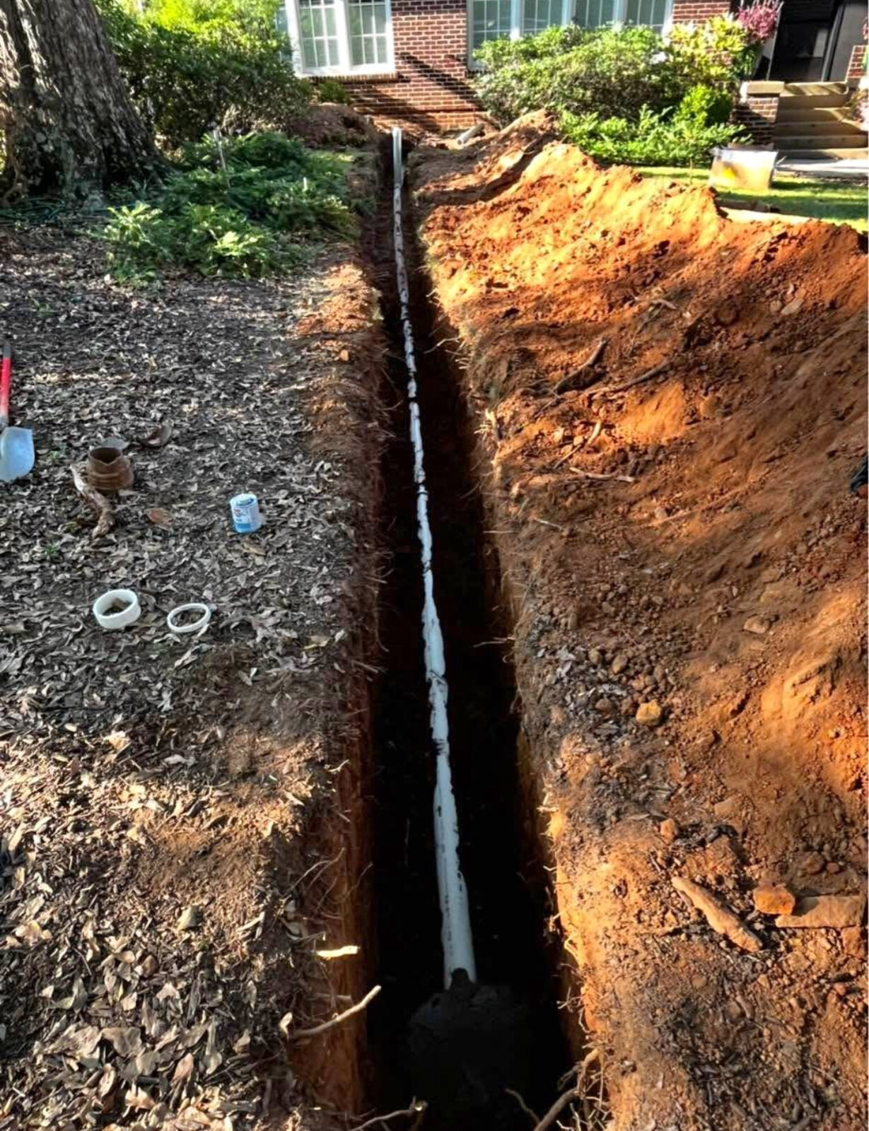 Open trench across a front yard with a new white PVC sewer line running toward the house